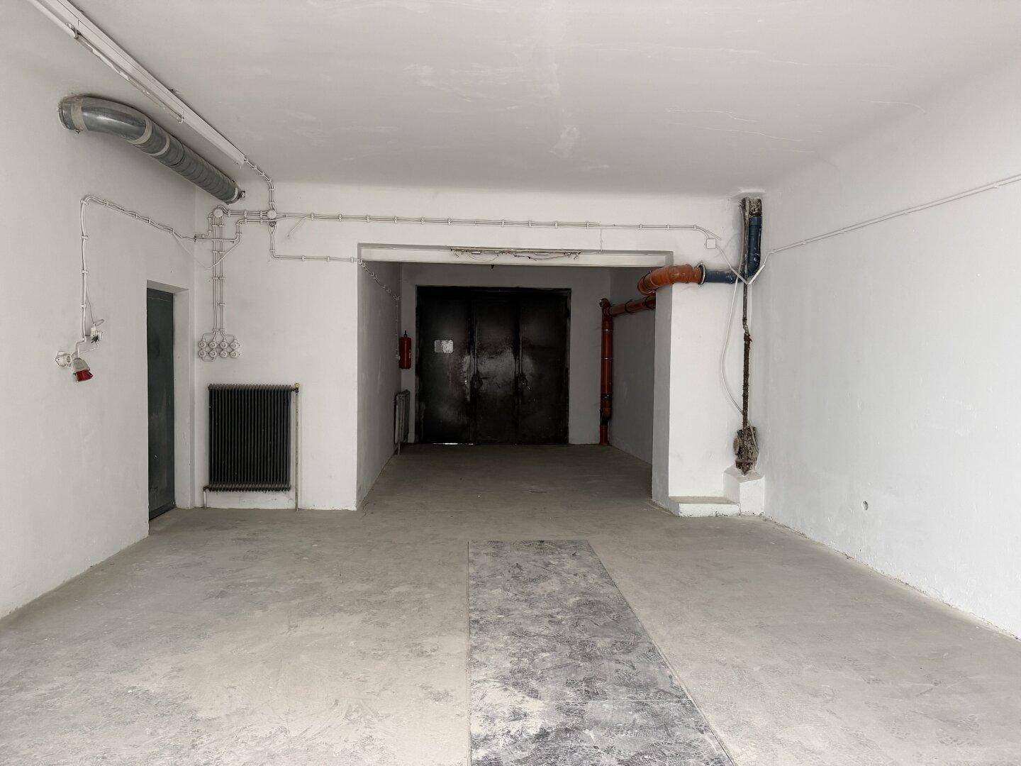 Großer Garagen- oder Lagerraum mit Betonboden, sichtbaren Rohrleitungen und robusten Doppeltüren im Hintergrund.