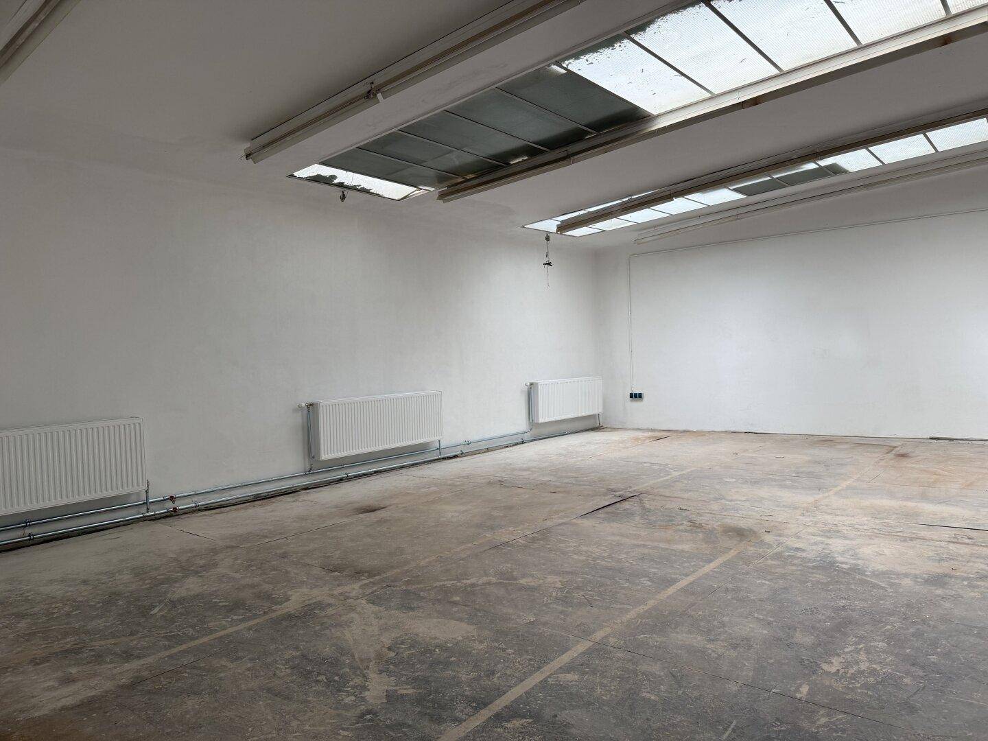 Leerer Gewerberaum mit weißen Wänden, Betonboden, zwei Heizkörpern und großen Oberlichtern, bereit zur Gestaltung.