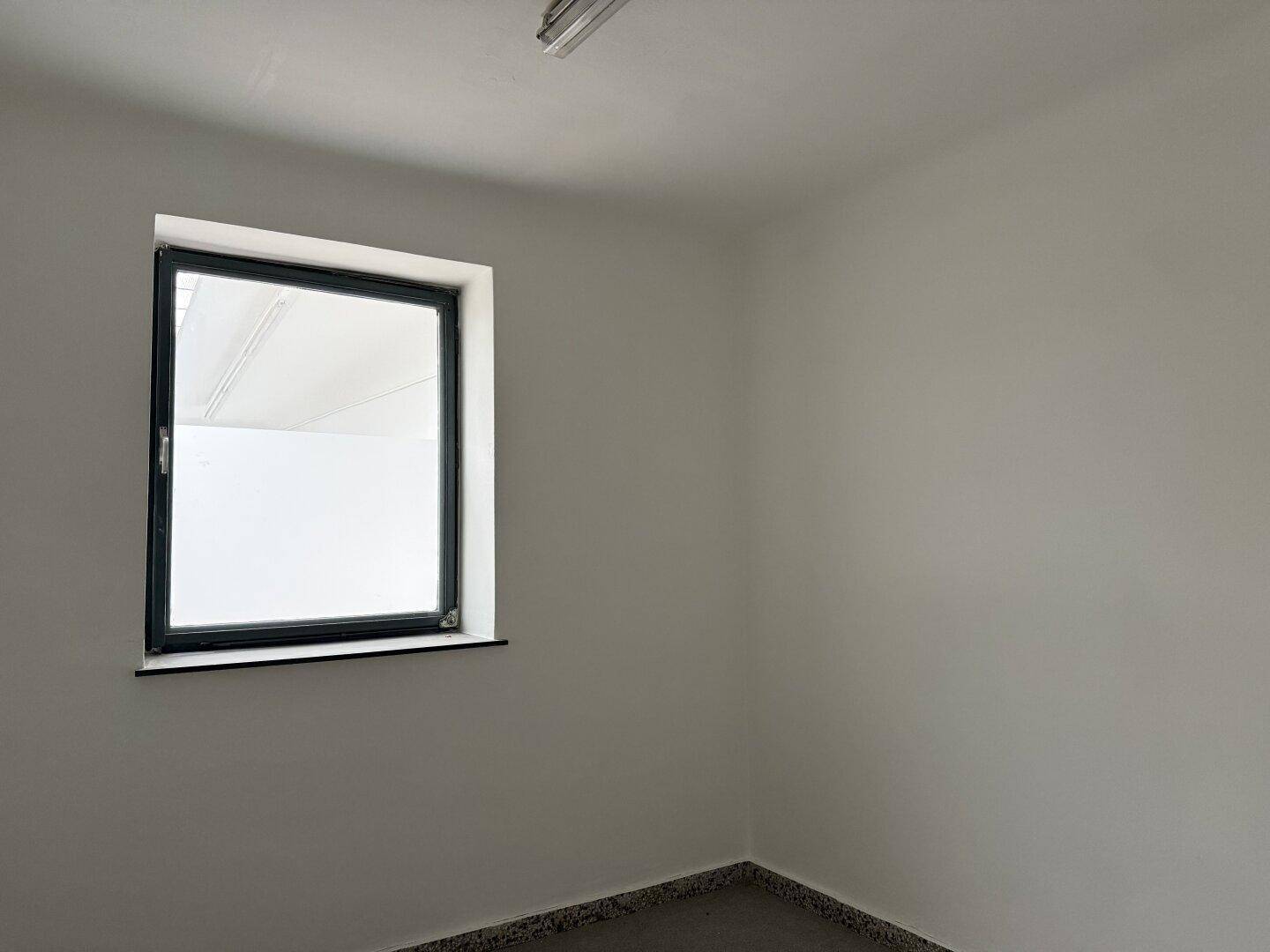 Kleiner, heller Raum mit weiß gestrichenen Wänden, einem Fenster und einem dunklen, gesprenkelten Fliesenboden.