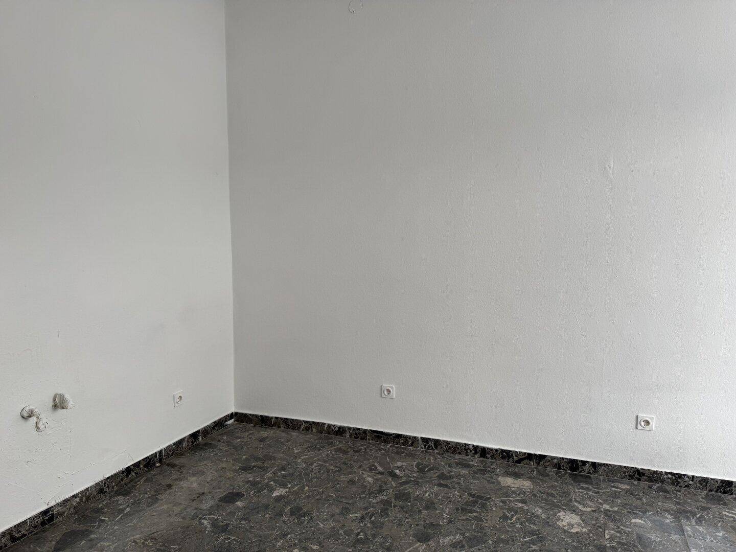 Ecke eines Raumes mit weißen Wänden und dunklem, gesprenkeltem Fliesenboden, ausgestattet mit Steckdosen.
