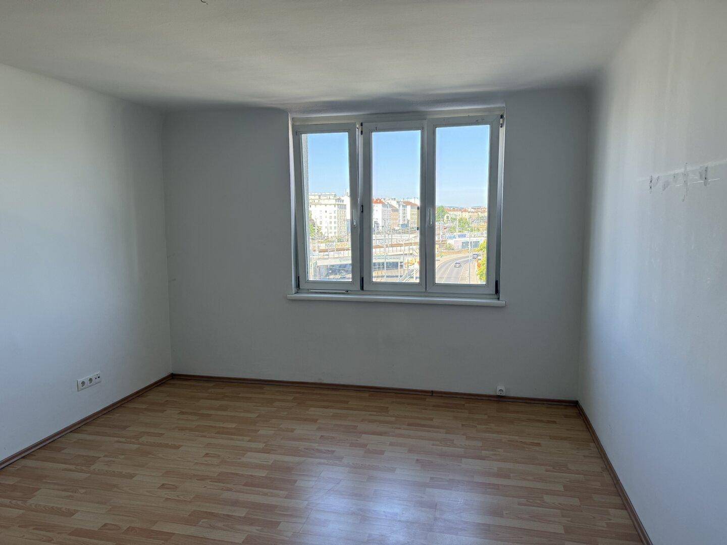 Heller, leerer Raum mit Laminatboden und großen Fenstern mit Stadtblick.
