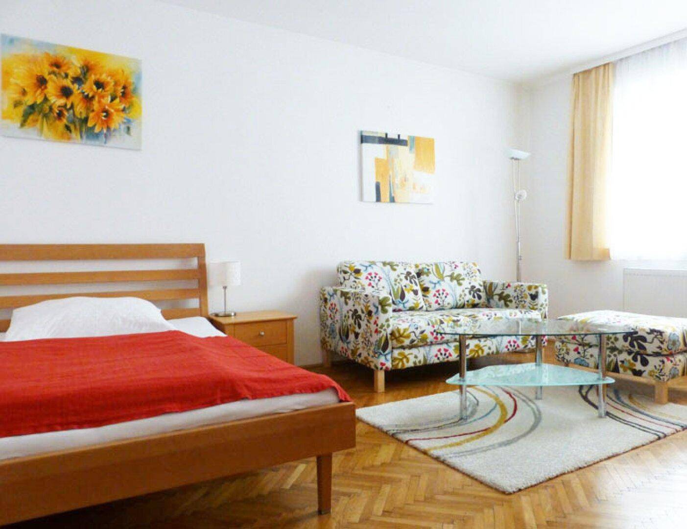 Gemütlicher Wohnbereich mit Bett, floralem Sofa, Teppich und viel Tageslicht durch das Fenster.