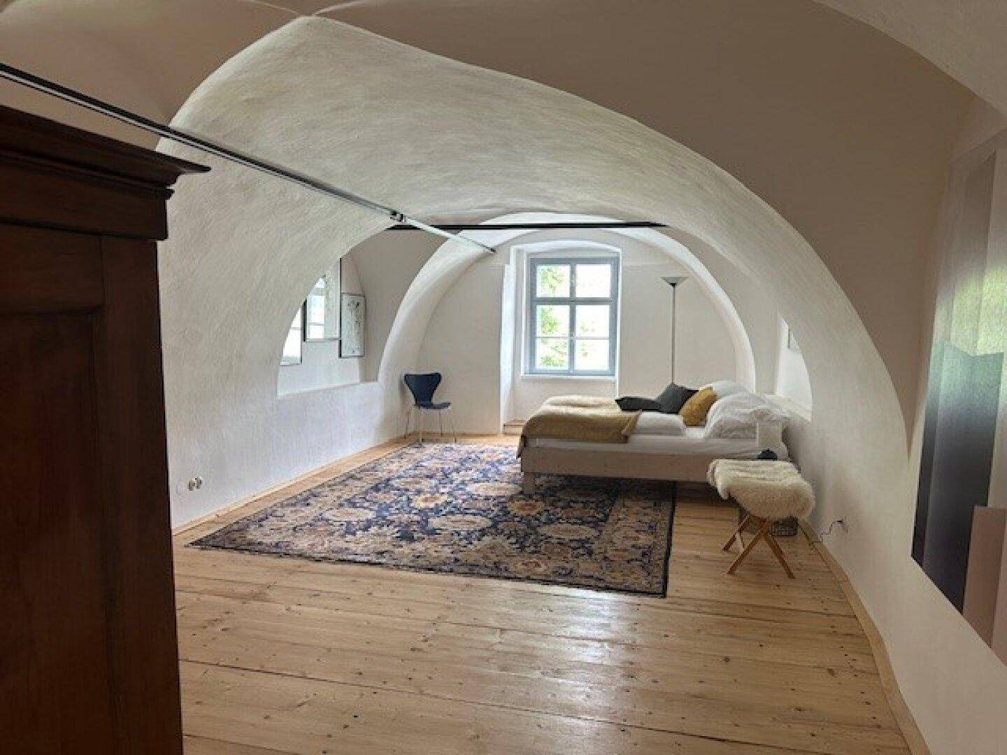 Helles Schlafzimmer mit gewölbter Decke, Holzdielenboden und einem großen Fenster, das viel Licht hereinlässt.