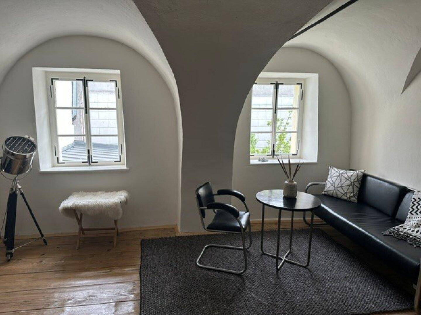 Gemütlicher Wohnbereich mit gewölbter Decke, zwei Fenstern und moderner schwarzer Sitzgelegenheit.