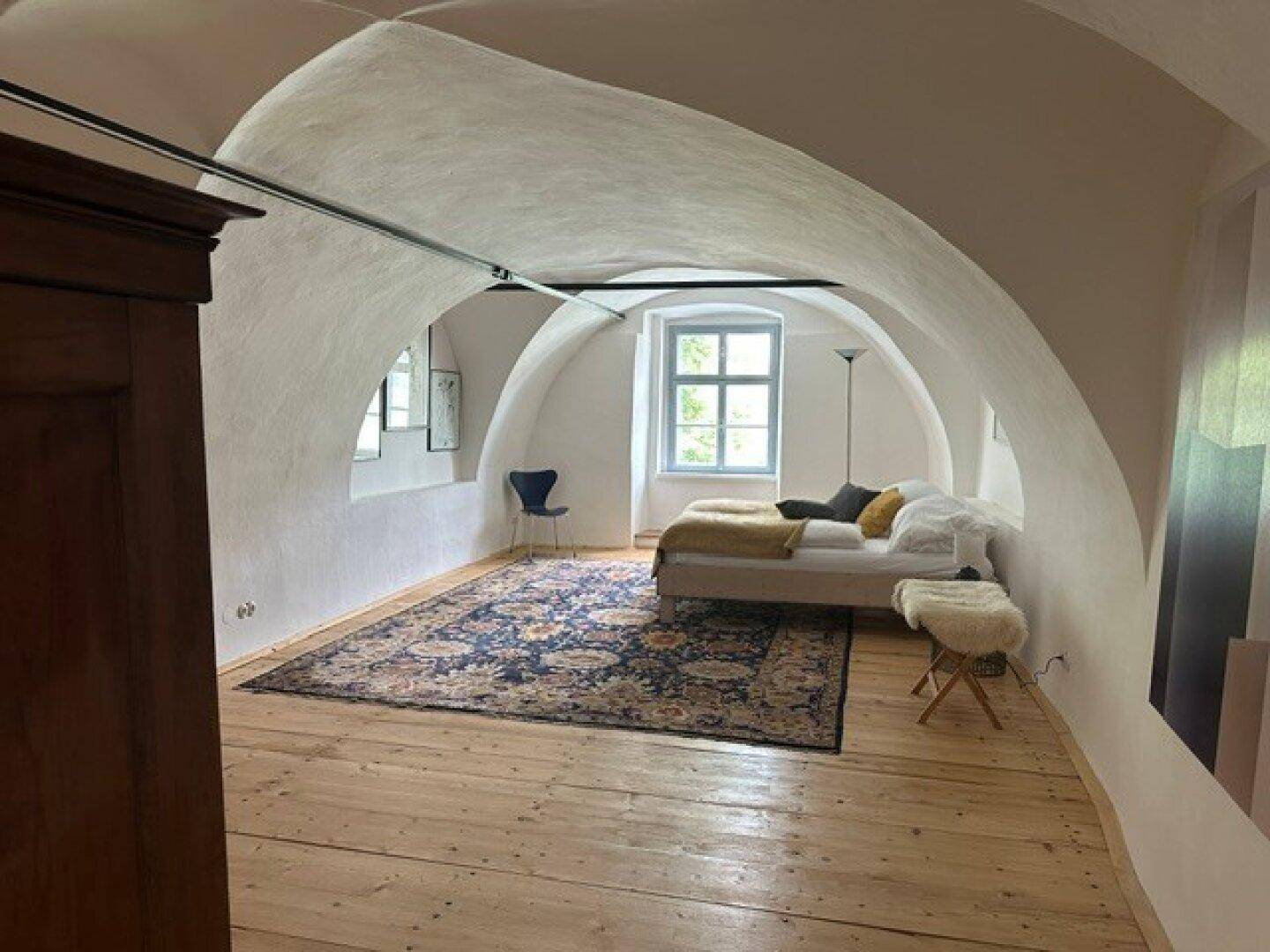 Helles Schlafzimmer mit gewölbter Decke, Holzdielenboden und einem großen Fenster, das viel Licht hereinlässt.