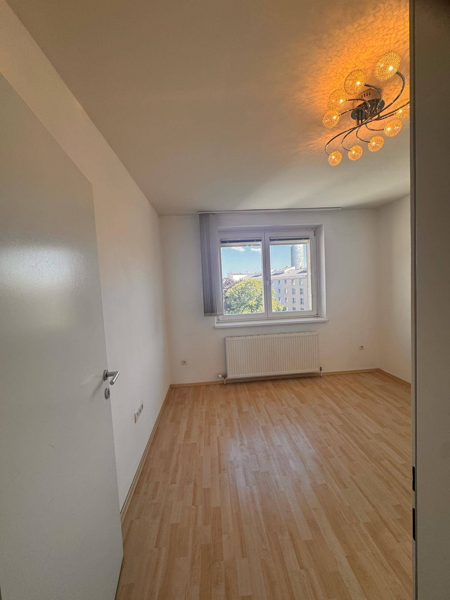 Geräumiges, helles Zimmer mit Laminatboden, großem Fenster und einer dekorativen Deckenleuchte.