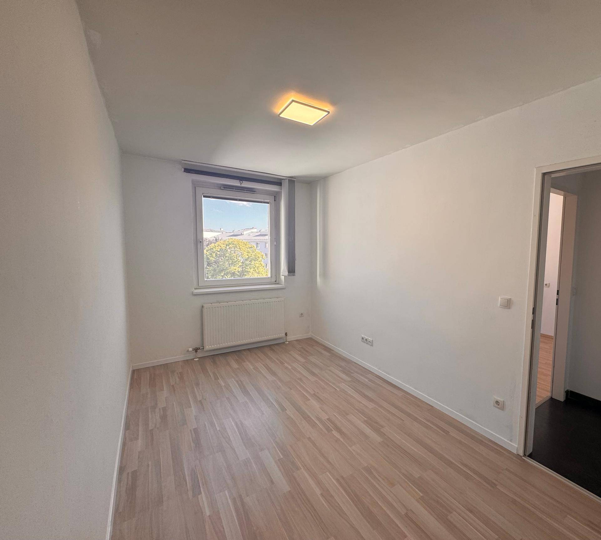 Helles, leeres Zimmer mit Laminatboden und einem Fenster, das viel Tageslicht hereinlässt.