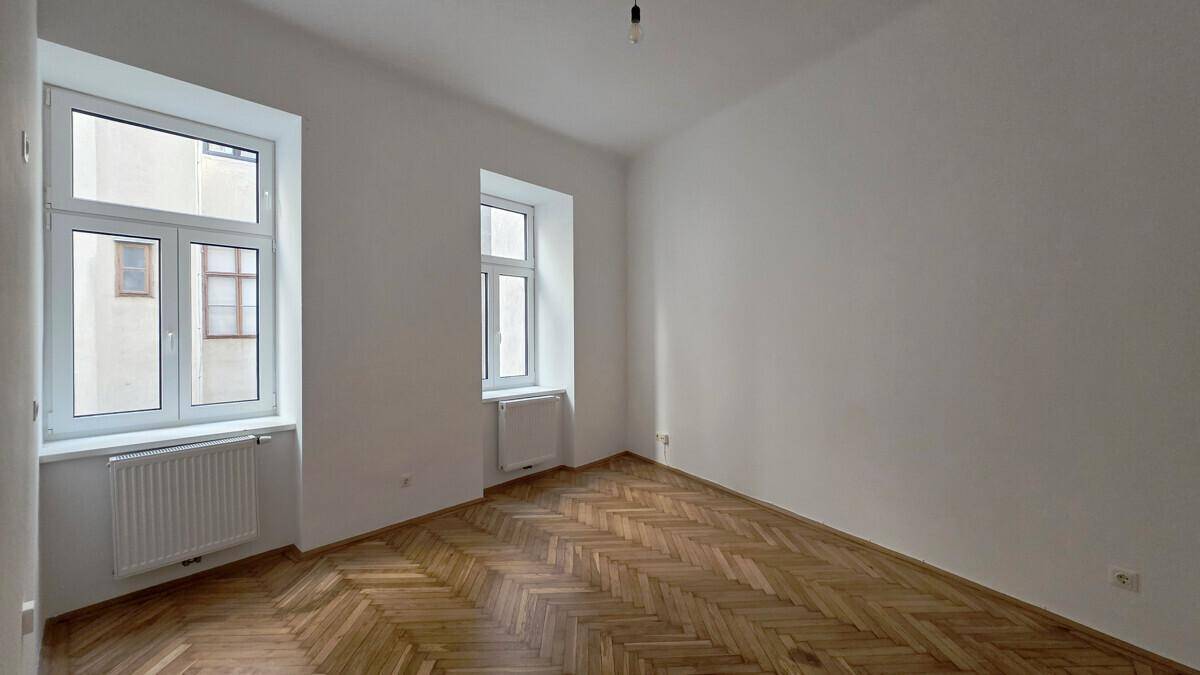 Geräumiges Zimmer mit zwei großen Fenstern und klassischem Fischgrätparkettboden.