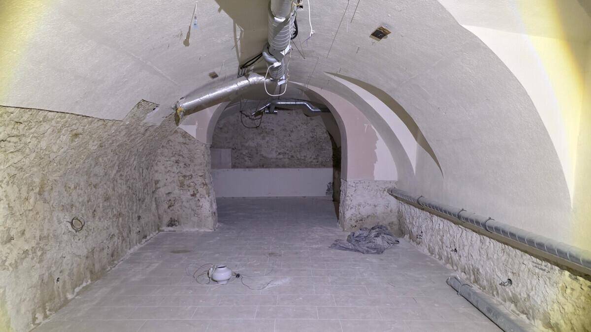 Langer Kellerraum mit Gewölbedecke, freigelegten Steinwänden und sichtbaren Lüftungsrohren, im Rohbauzustand.