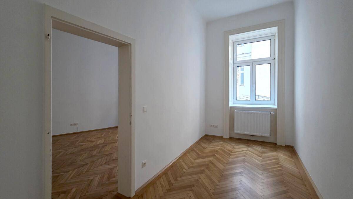 Lichtdurchfluteter Raum mit Fischgrätparkett und Zugang zu einem weiteren Zimmer.