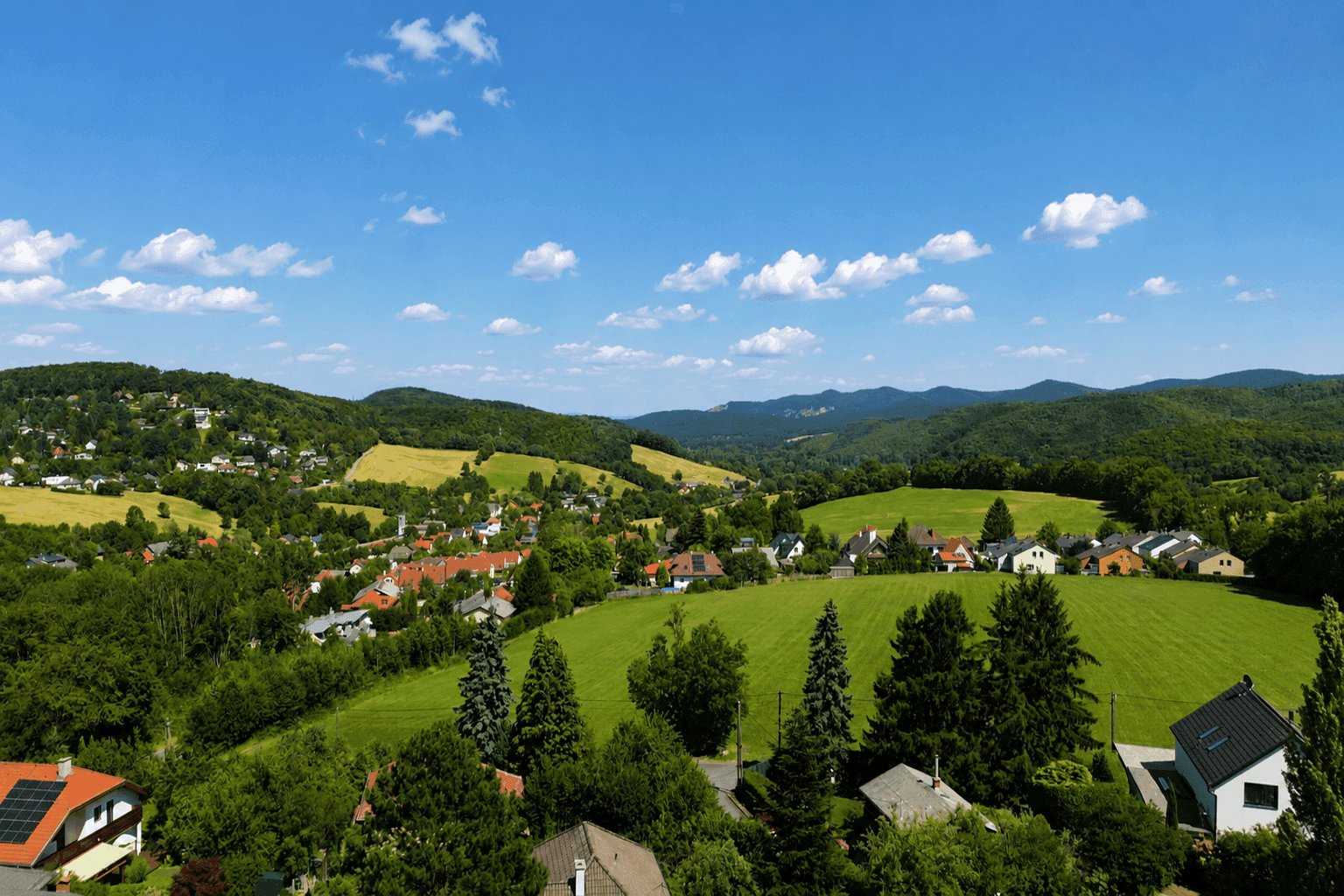 Weitläufige Hügellandschaft mit grünen Feldern, Wäldern und verstreuten Häusern unter blauem Himmel.