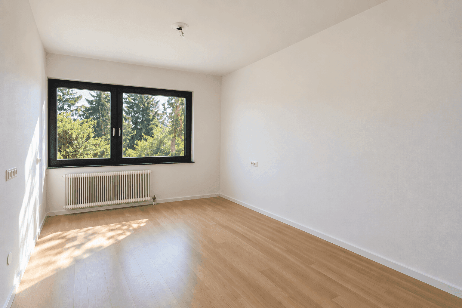 Leeres Zimmer mit Holzboden und großem Fenster, das viel natürliches Licht hereinlässt.
