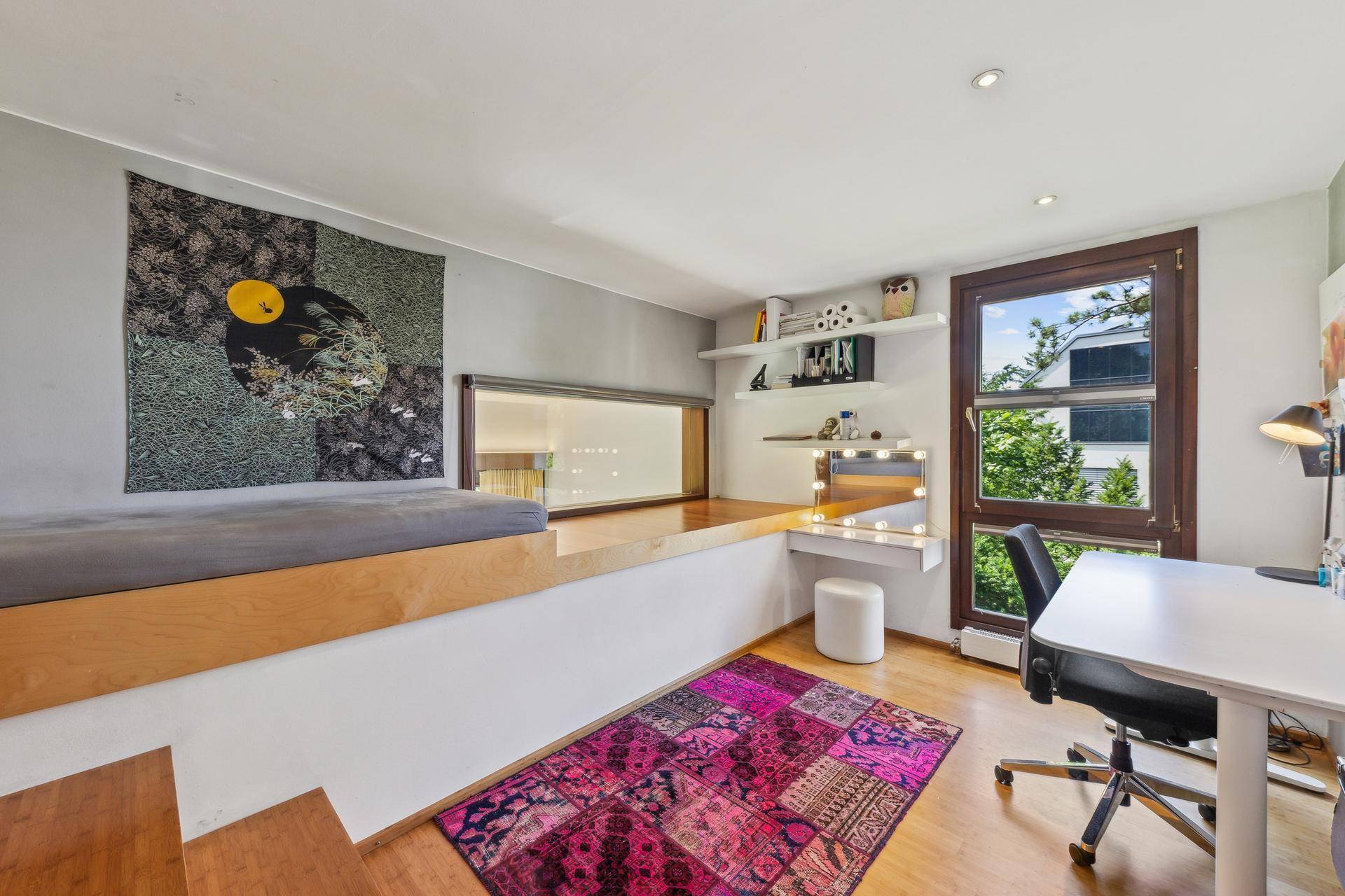 Helles Zimmer mit erhöhtem Schlafbereich, Schreibtisch und farbenfrohem Teppich, ideal für Kinder oder Gäste.