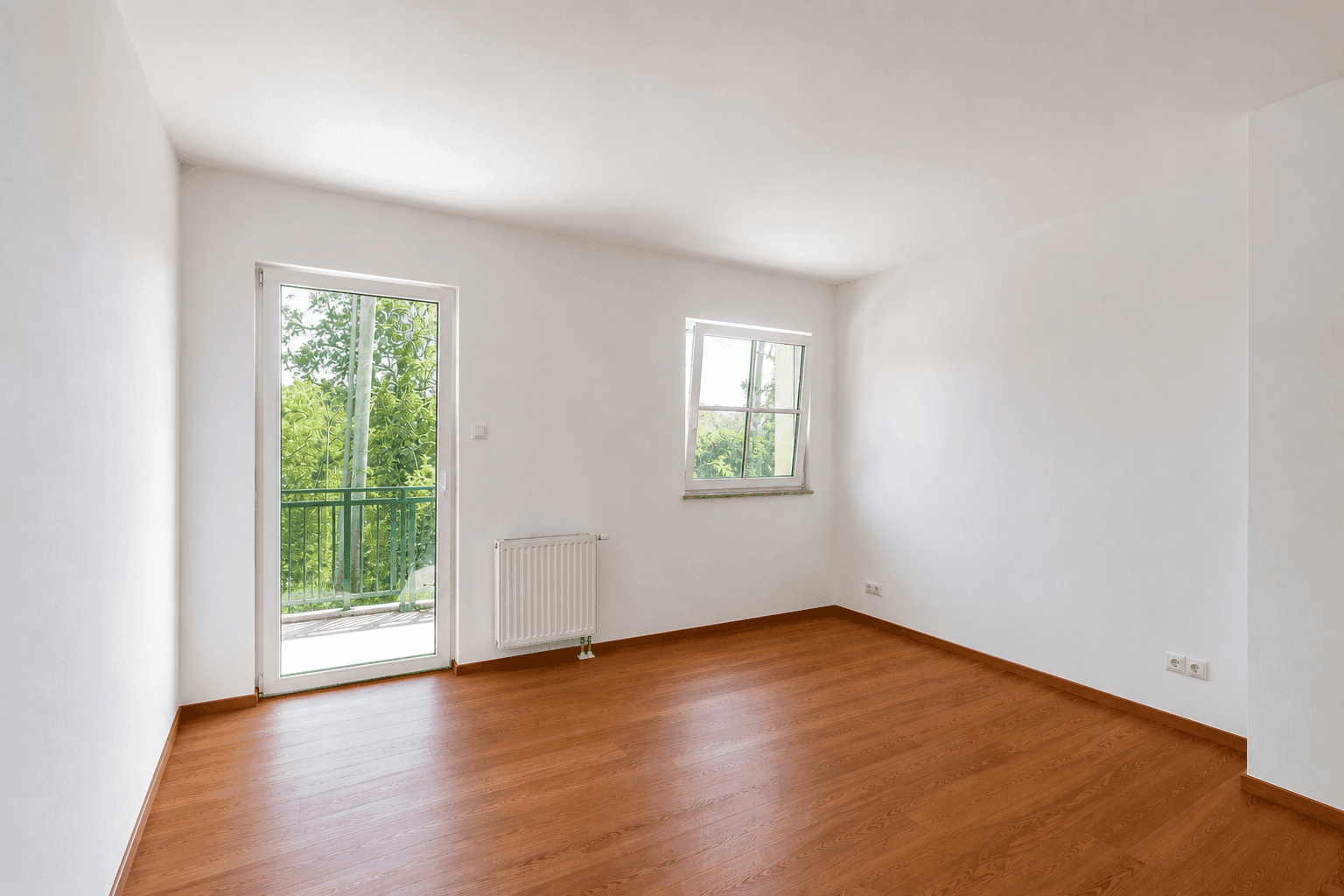 Leeres Schlafzimmer mit Holzboden, Heizkörper und Zugang zu einem Balkon.