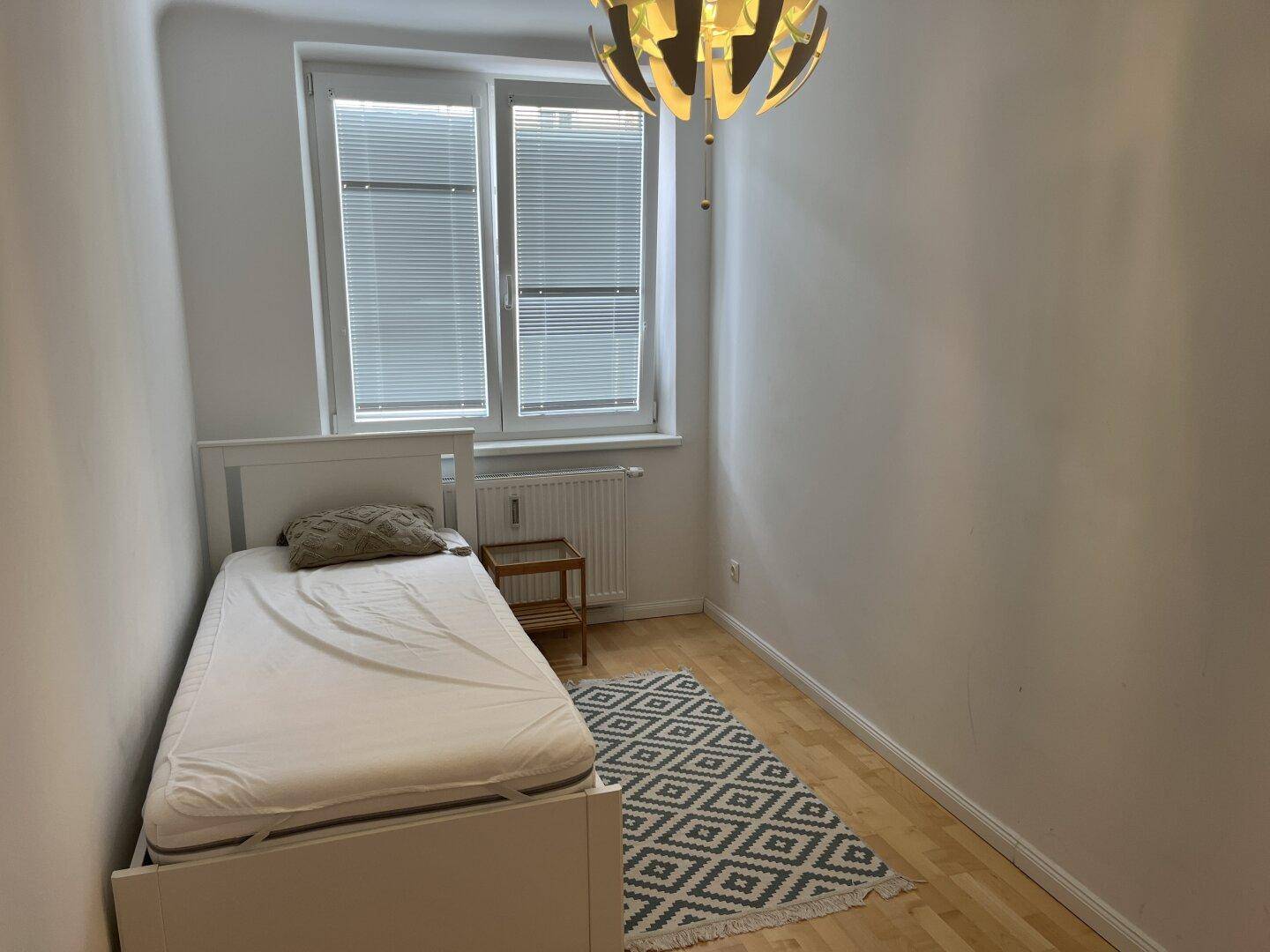 Helles Schlafzimmer mit Einzelbett, Fenster und Teppich auf Holzboden.