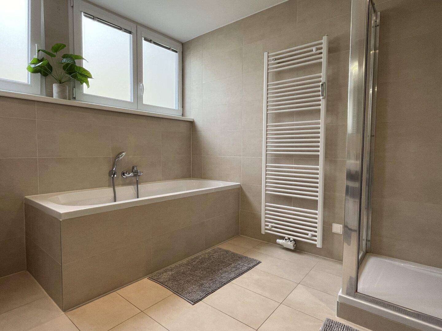 Helles Badezimmer mit eingebauter Badewanne, Fenster und modernem Handtuchheizkörper.