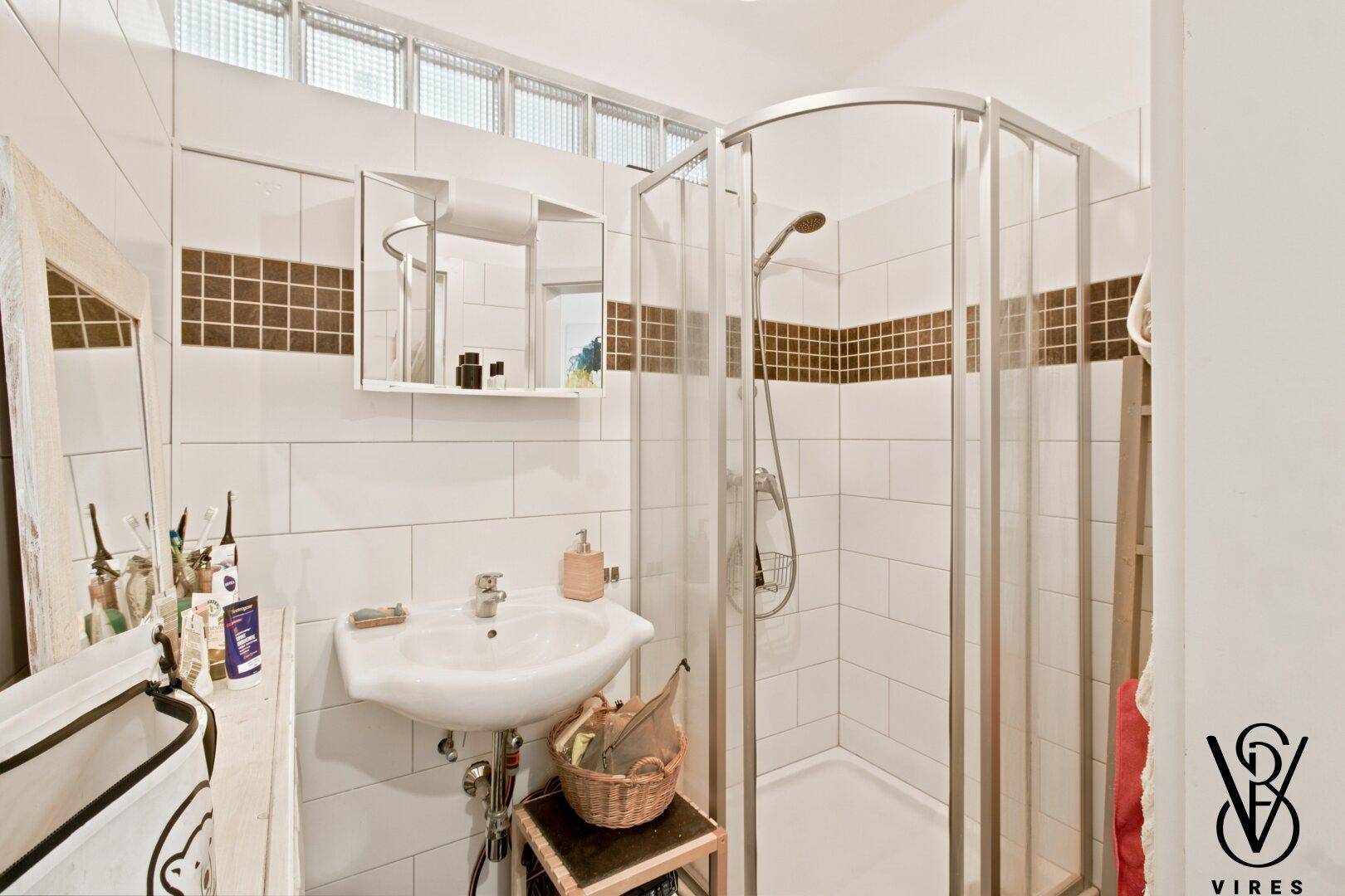 Badezimmer mit Duschkabine, Waschbecken und weißen Fliesen mit braunem Akzentstreifen.