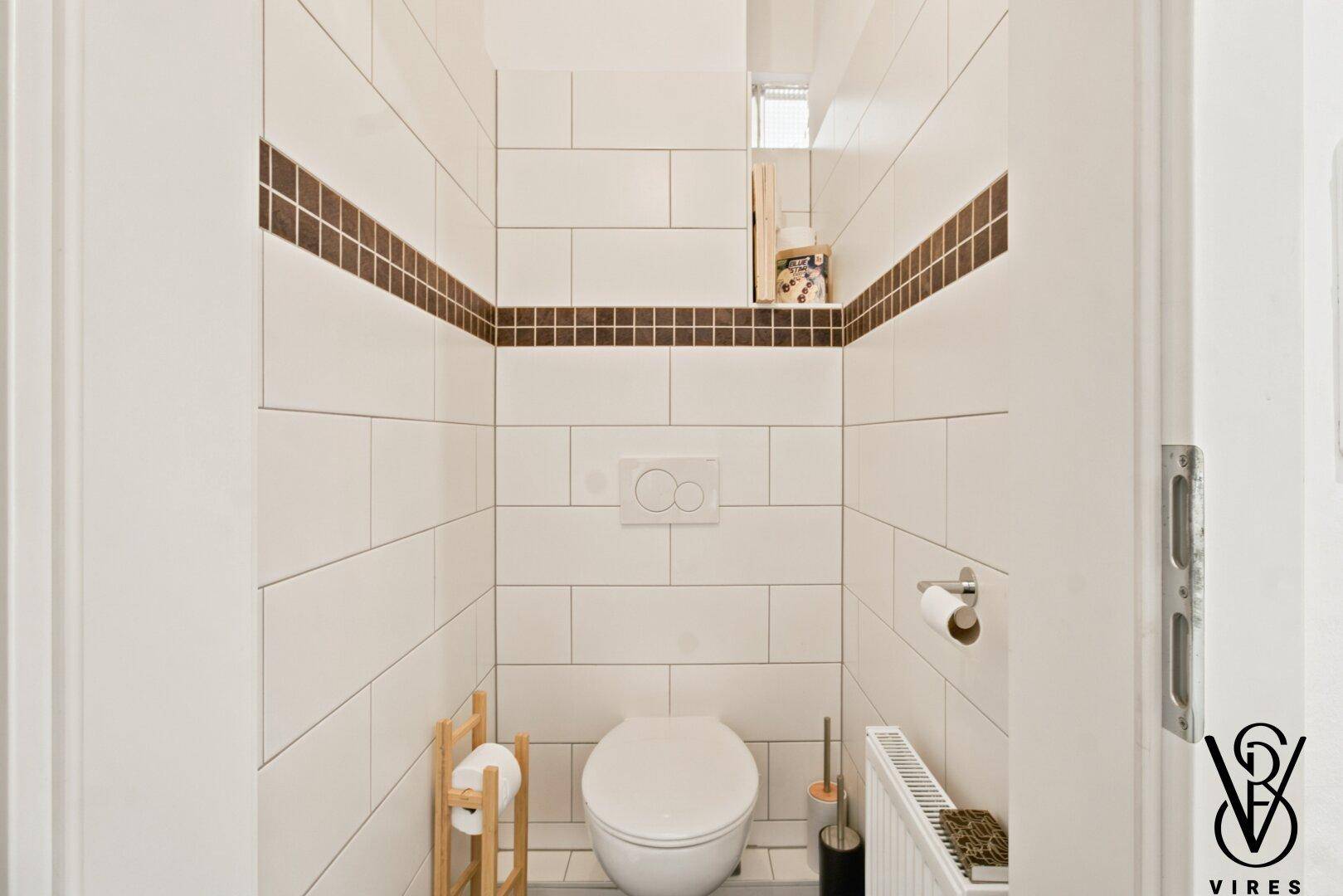 Separate Toilette mit wandhängendem WC und weißen Fliesen mit braunem Akzentstreifen.