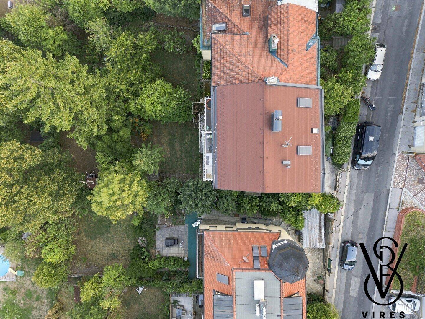 Draufsicht auf zwei Wohnhäuser mit Gärten und einer angrenzenden Straße, umgeben von Bäumen.