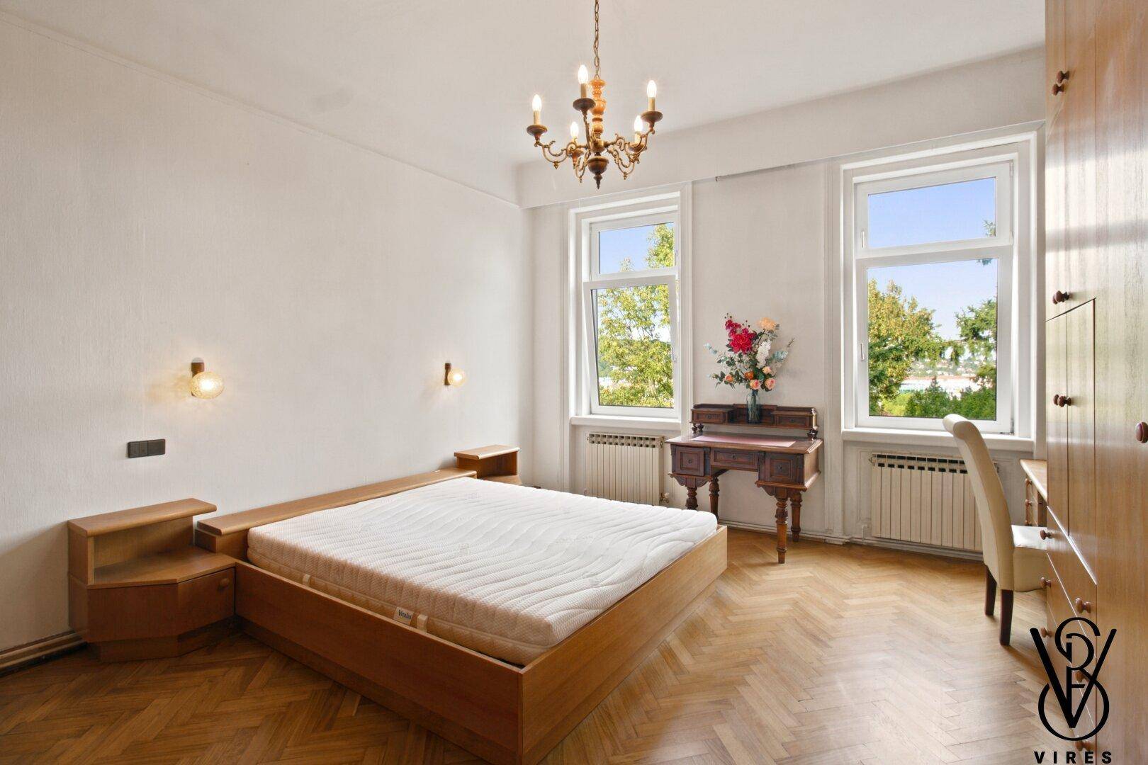 Schlafzimmer mit Fischgrätparkett, Doppelbett und großen Fenstern mit Blick ins Grüne.