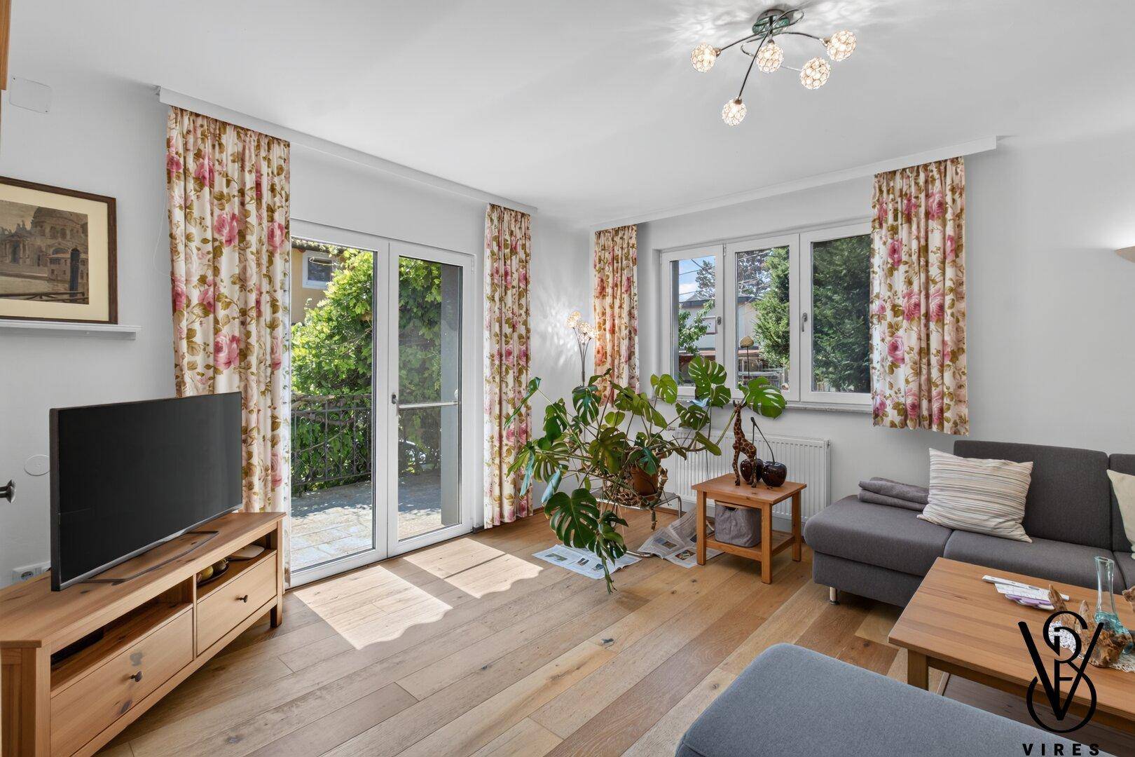 Helles Wohnzimmer mit Zugang zur Terrasse, Fernseher und Blick ins Grüne durch große Fenster.