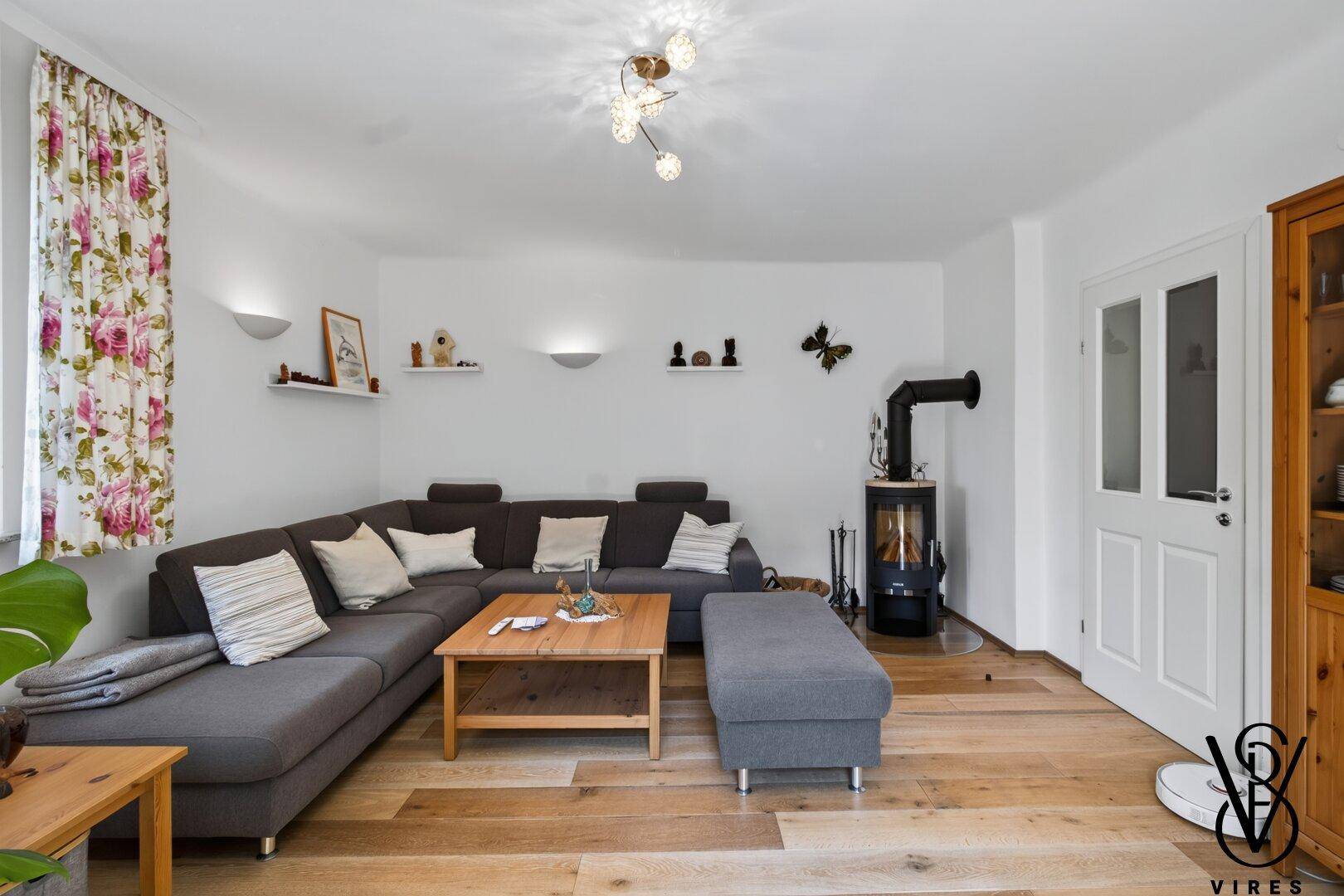 Gemütliches Wohnzimmer mit großem Ecksofa, Holzboden und modernem Holzofen als Wärmequelle.