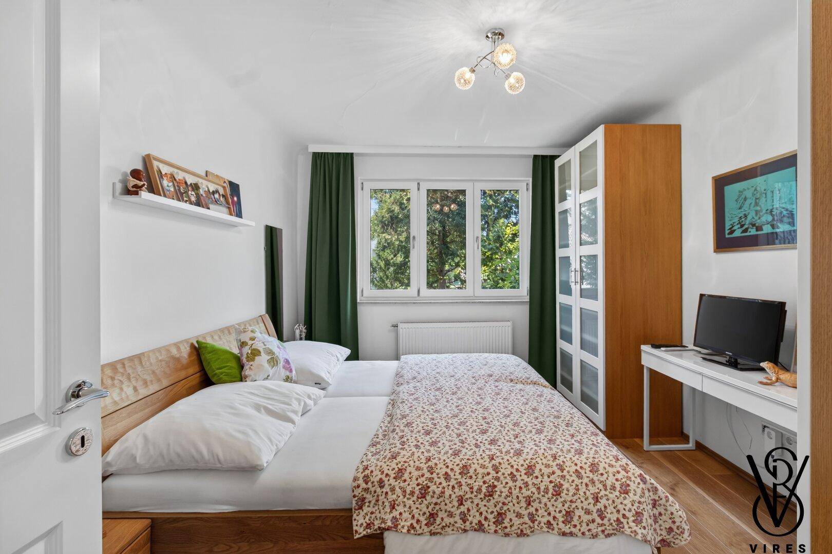 Schlafzimmer mit Blick ins Grüne, Holzbett und Schreibtisch am Fenster.