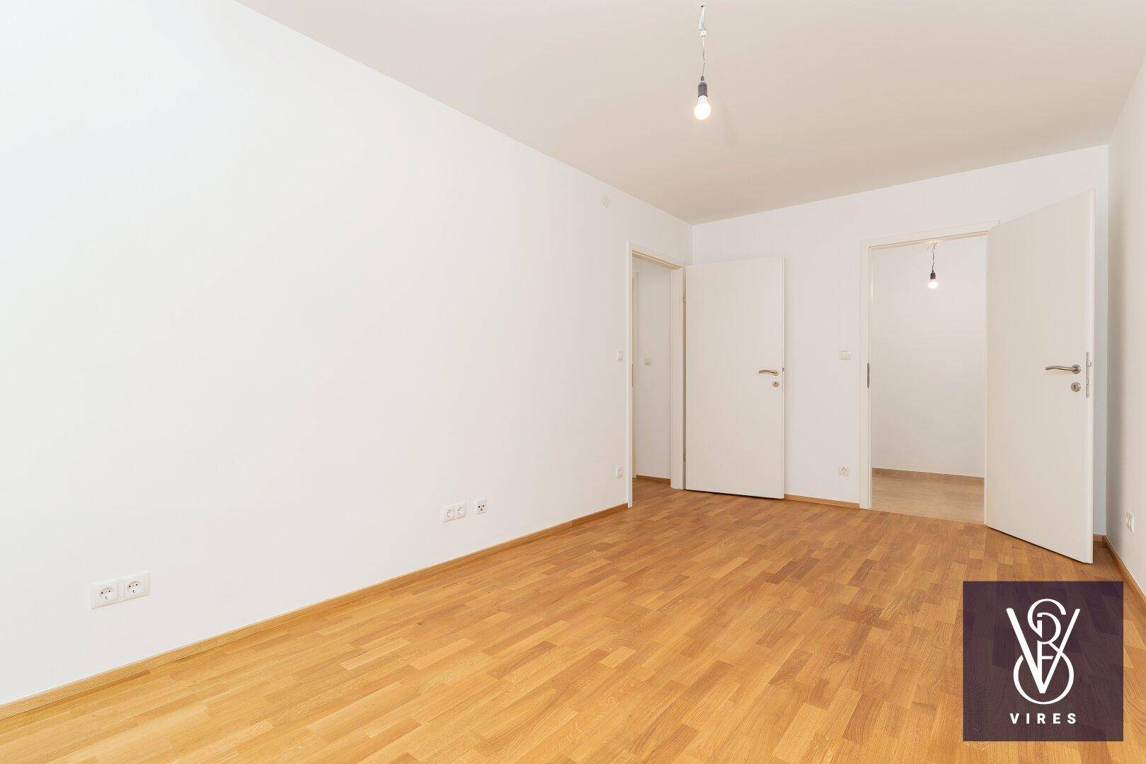 Leeres Schlafzimmer mit Parkettboden und zwei Türen, die zu weiteren Räumen führen.