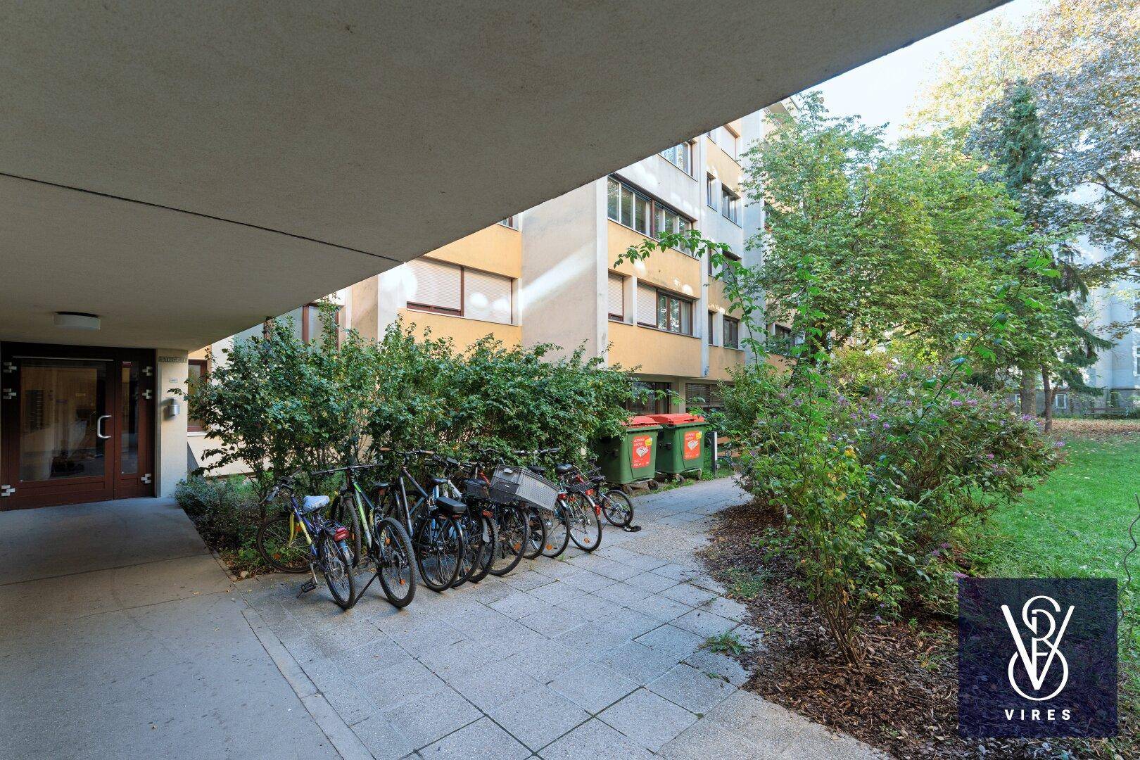 Eingangsbereich eines Gebäudes mit Fahrradstellplätzen und grüner Bepflanzung.