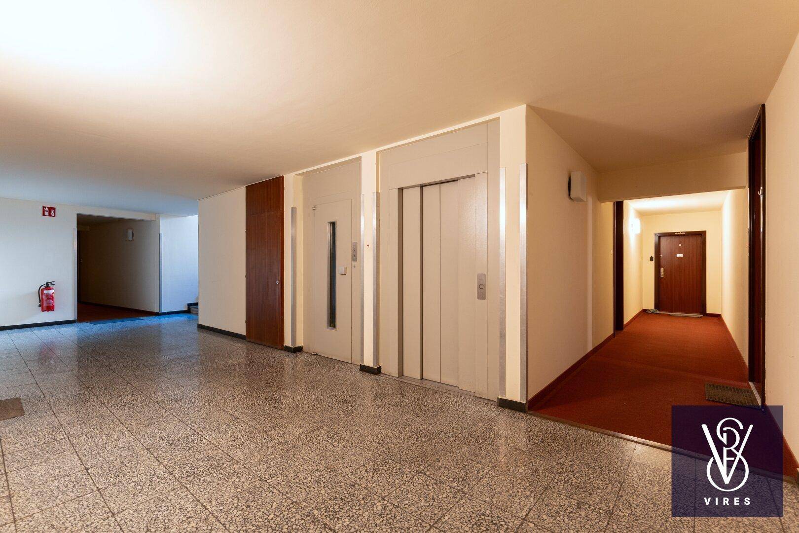 Geräumiger Flur im Gebäude mit Fliesenboden, mehreren Türen und einem Aufzug.