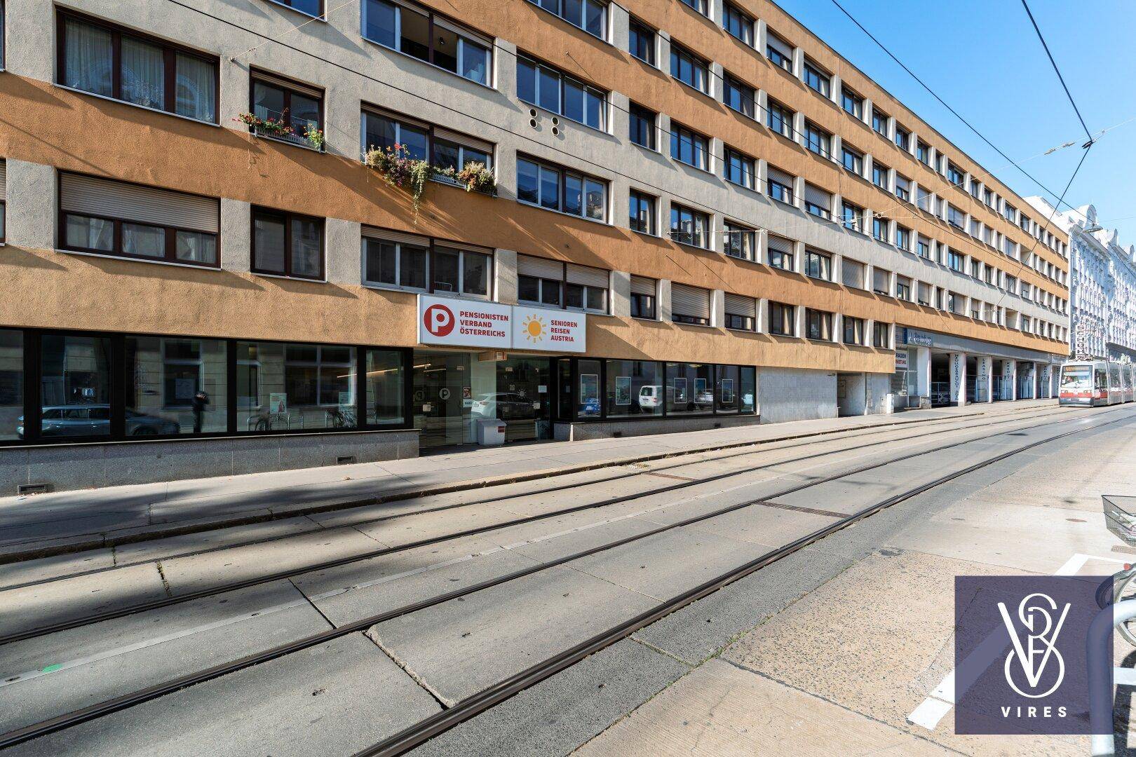Mehrstöckiges Gebäude mit Geschäften im Erdgeschoss und Straßenbahnschienen im Vordergrund.