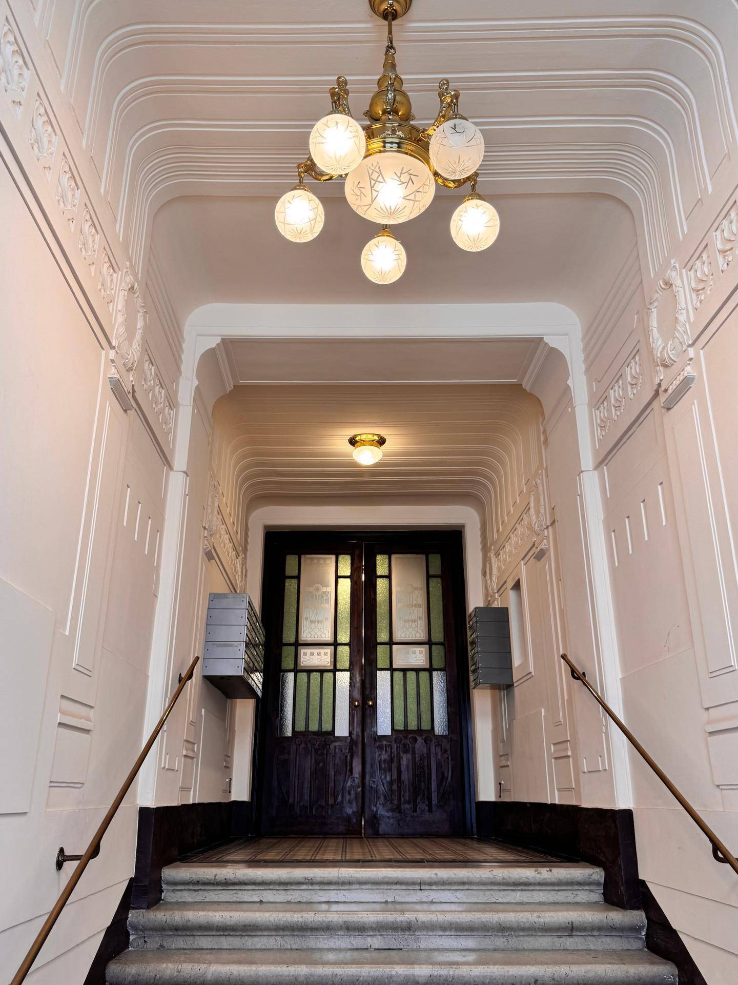 Elegantes Treppenhaus mit Stuckdecken, prunkvollem Kronleuchter und historischen Eingangstüren.