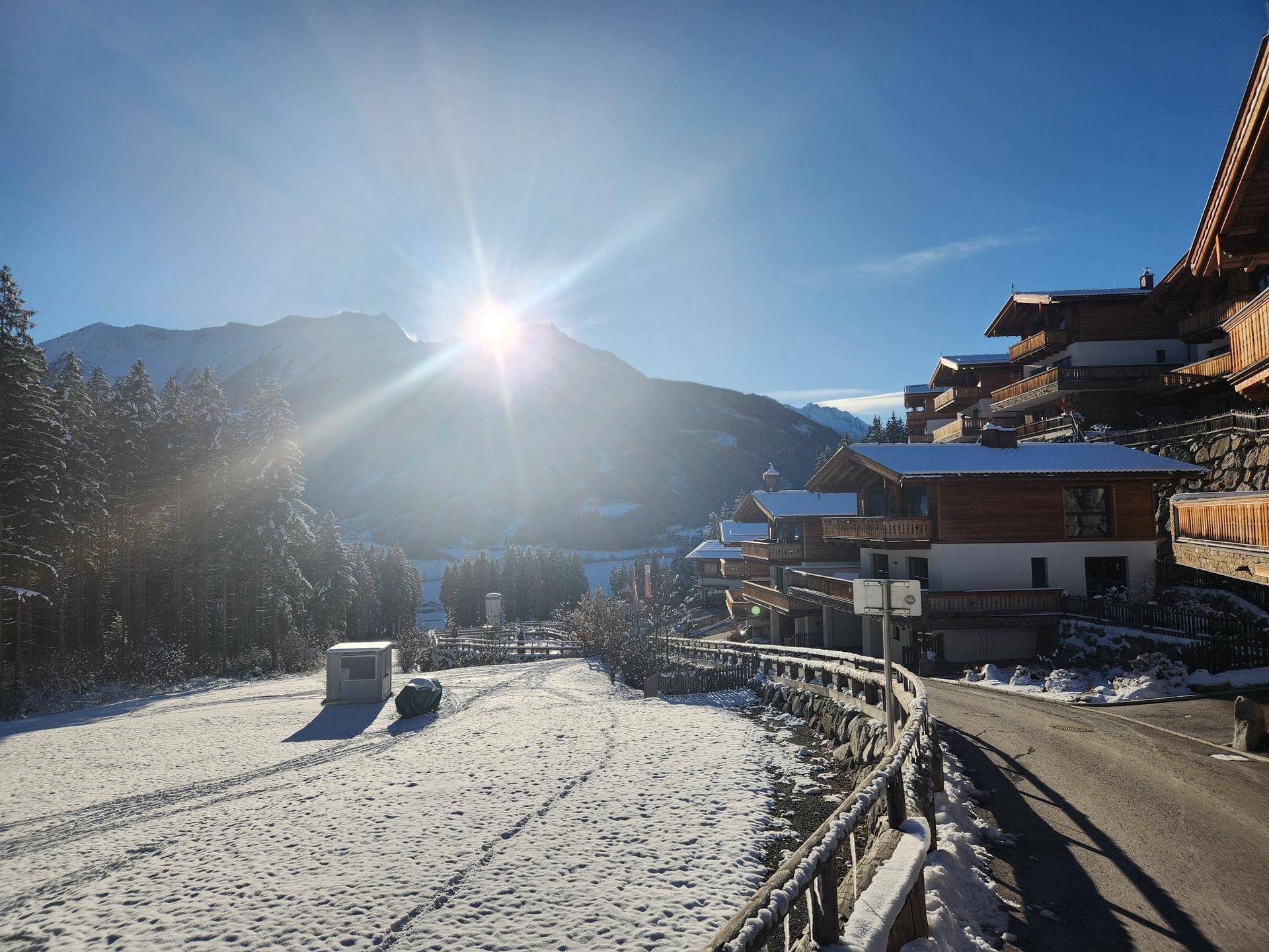Winterliche Landschaft mit Chalet-Siedlung, verschneiten Feldern und Bergen im Hintergrund.