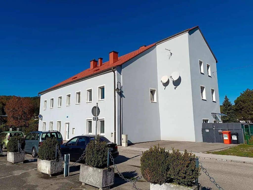 Gebäudeseite mit hellblauer Fassade, Satellitenschüsseln und Mülltonnen im Außenbereich.