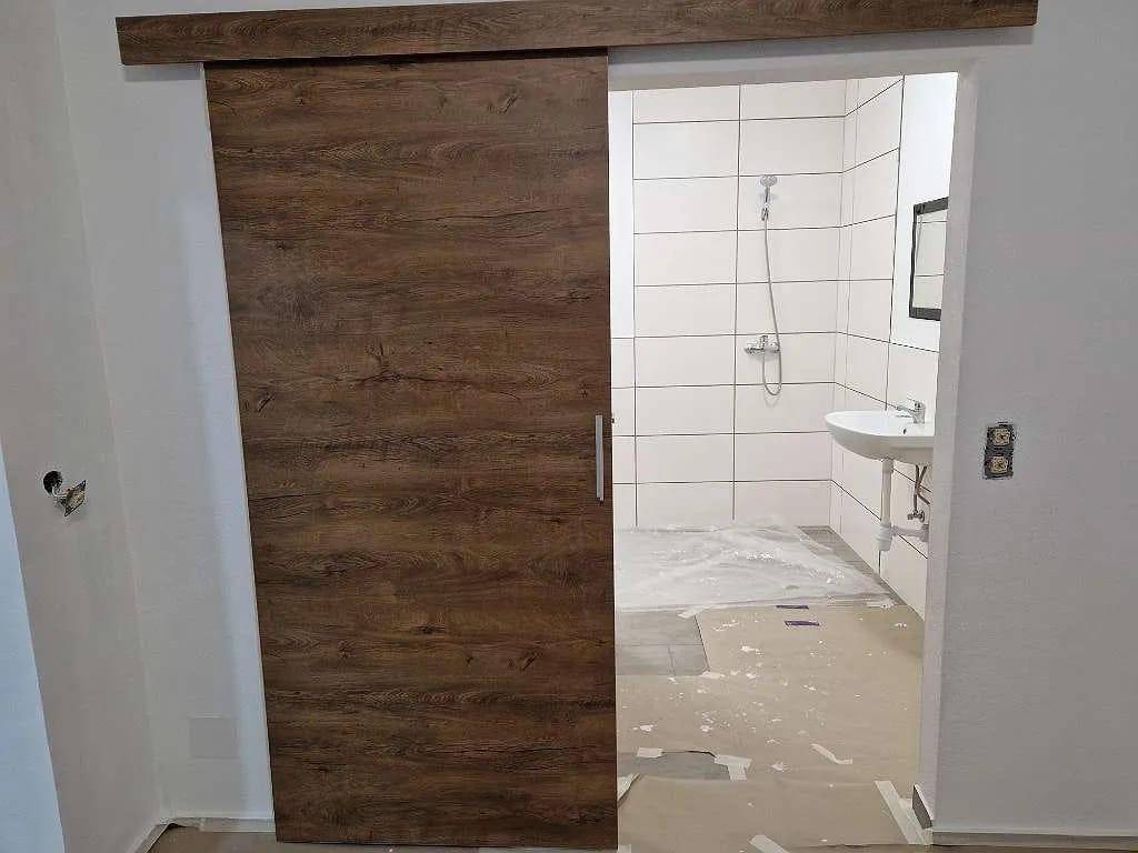 Badezimmer im Bau mit weißen Fliesen, Waschbecken und Duschanschluss hinter einer Schiebetür.