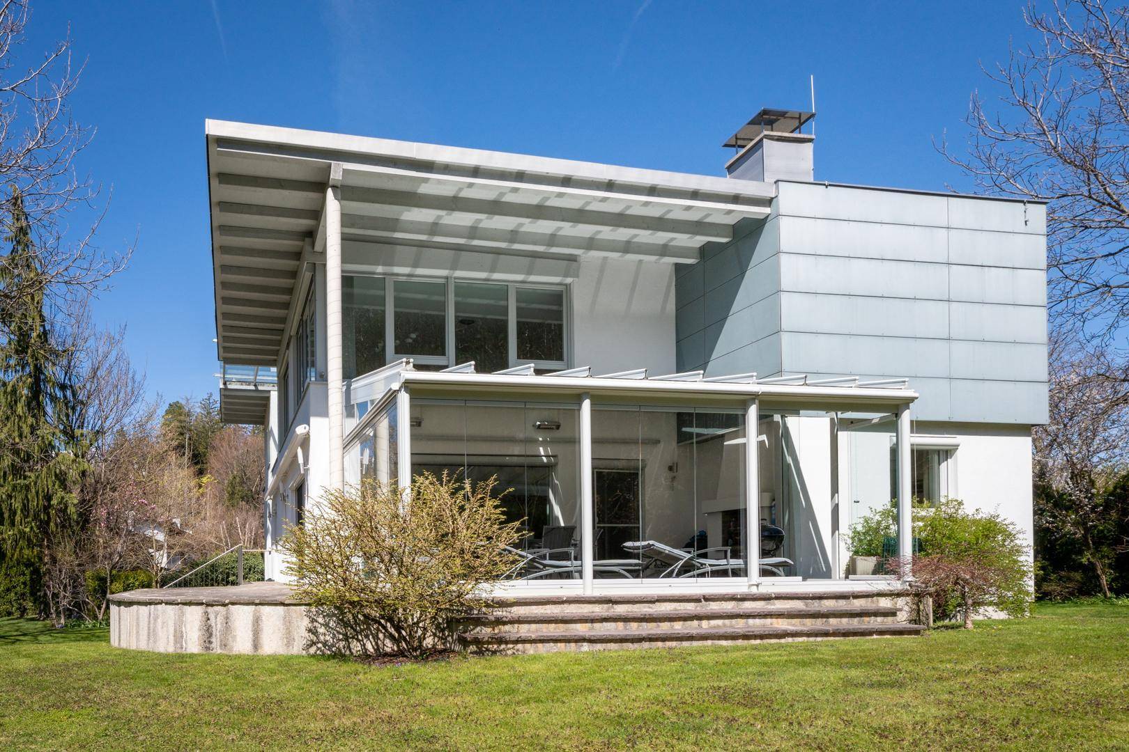 Rückansicht des Hauses mit verglastem Wintergarten und einer sonnigen Terrasse, umgeben von Grünflächen.