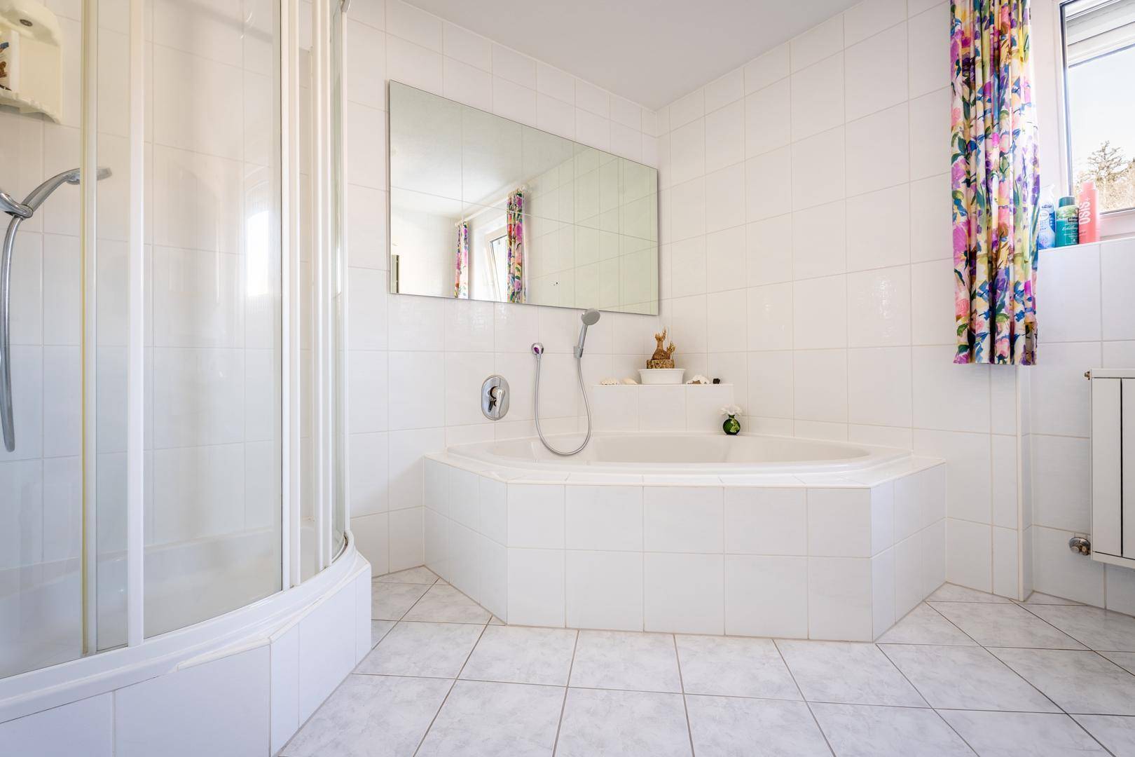 Badezimmer mit Eckbadewanne, Duschkabine und weißen Fliesen, ausgestattet mit bunten Vorhängen.