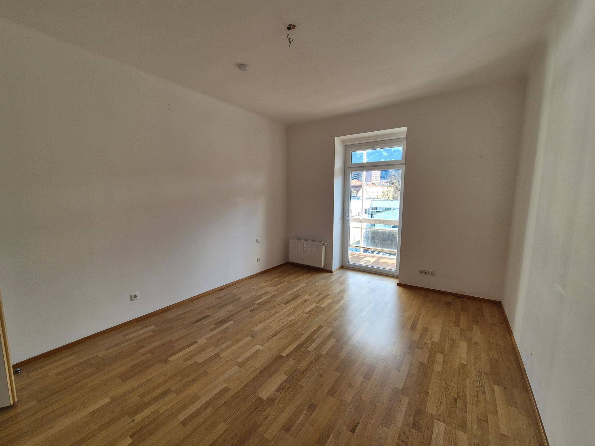 Heller, leerer Raum mit Holzboden und großem Fenster, das viel Tageslicht hereinlässt.