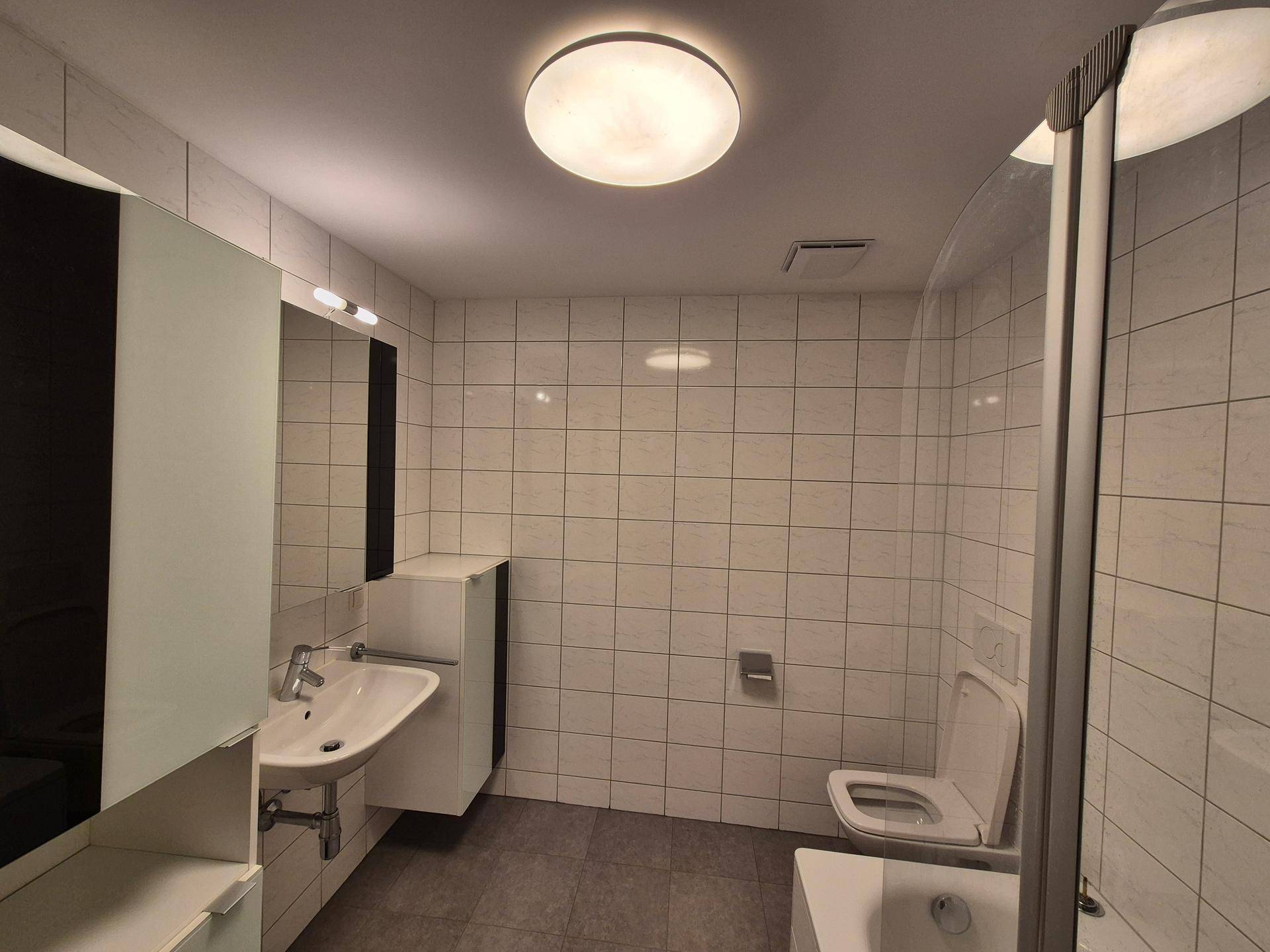 Modernes Badezimmer mit weißen Fliesen, Duschkabine und wandhängendem WC.