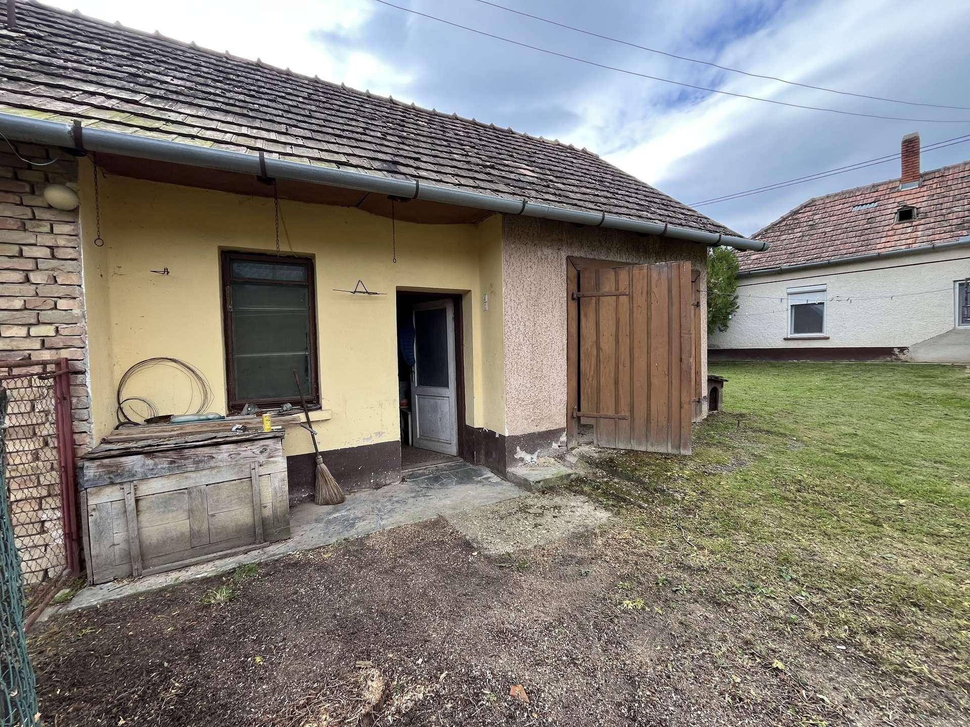 Außenansicht eines kleinen Gebäudes mit gelber Fassade, Holztür und einem einfachen gepflasterten Bereich.