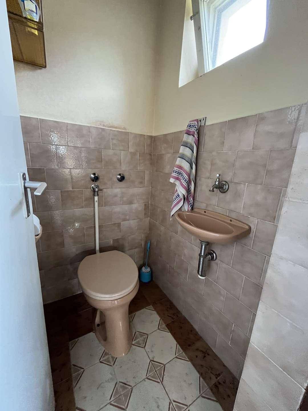 Kleines, älteres WC mit beigefarbenen Fliesen, einem kleinen Waschbecken und einem Fenster.
