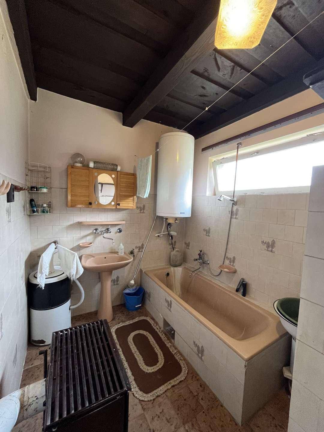 Älteres Badezimmer mit Badewanne, Waschbecken, Warmwasserboiler und weißen Fliesen.