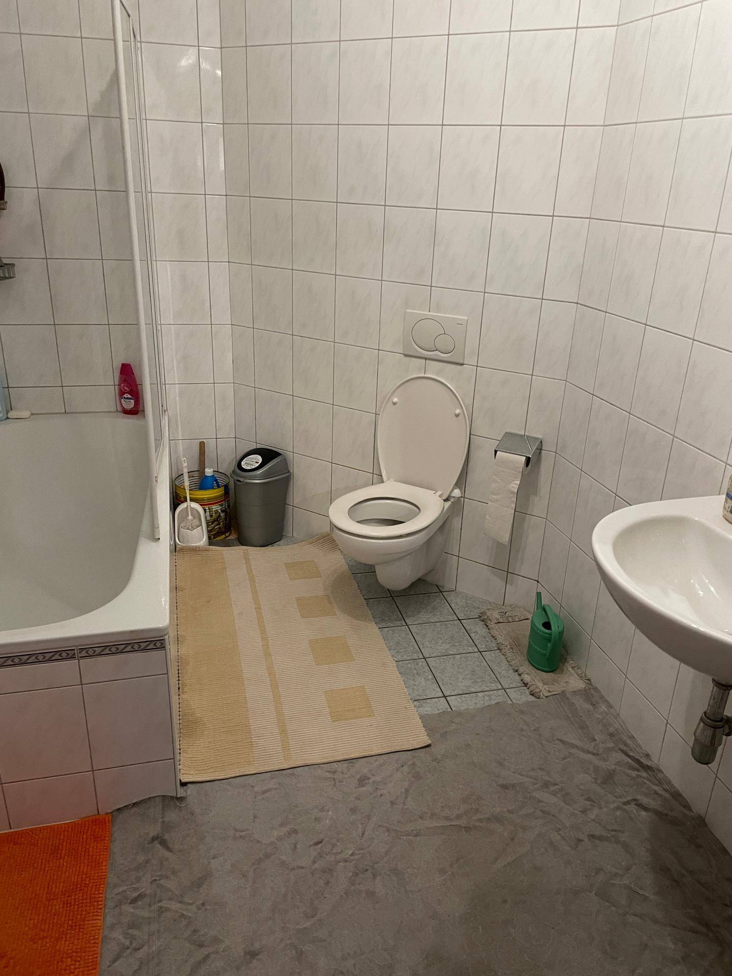 Badezimmer mit Toilette, Waschbecken und Badewanne, ausgestattet mit weißen Fliesen und einem Teppich.