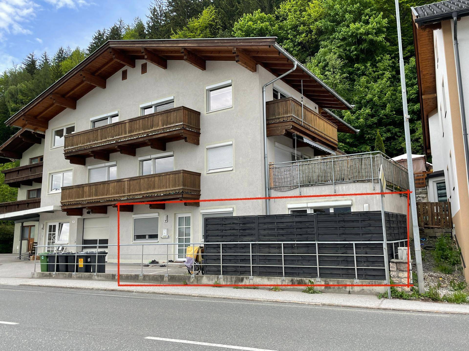 Gebäudefassade mit Holzbalkonen und einem eingezäunten Bereich im Erdgeschoss, umgeben von Grün.