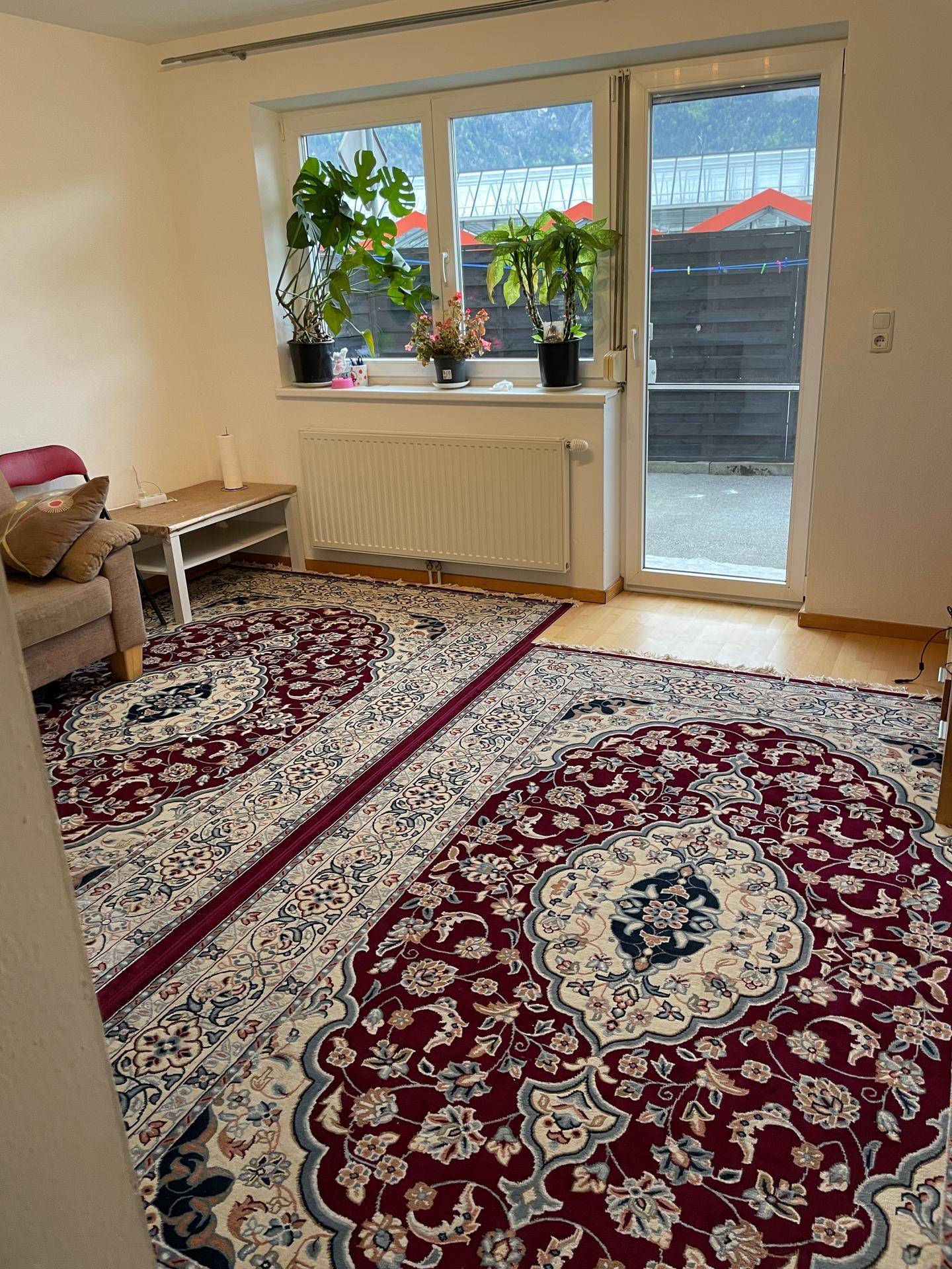 Wohnzimmer mit großen Fenstern, traditionellen Teppichen und Pflanzen auf der Fensterbank.