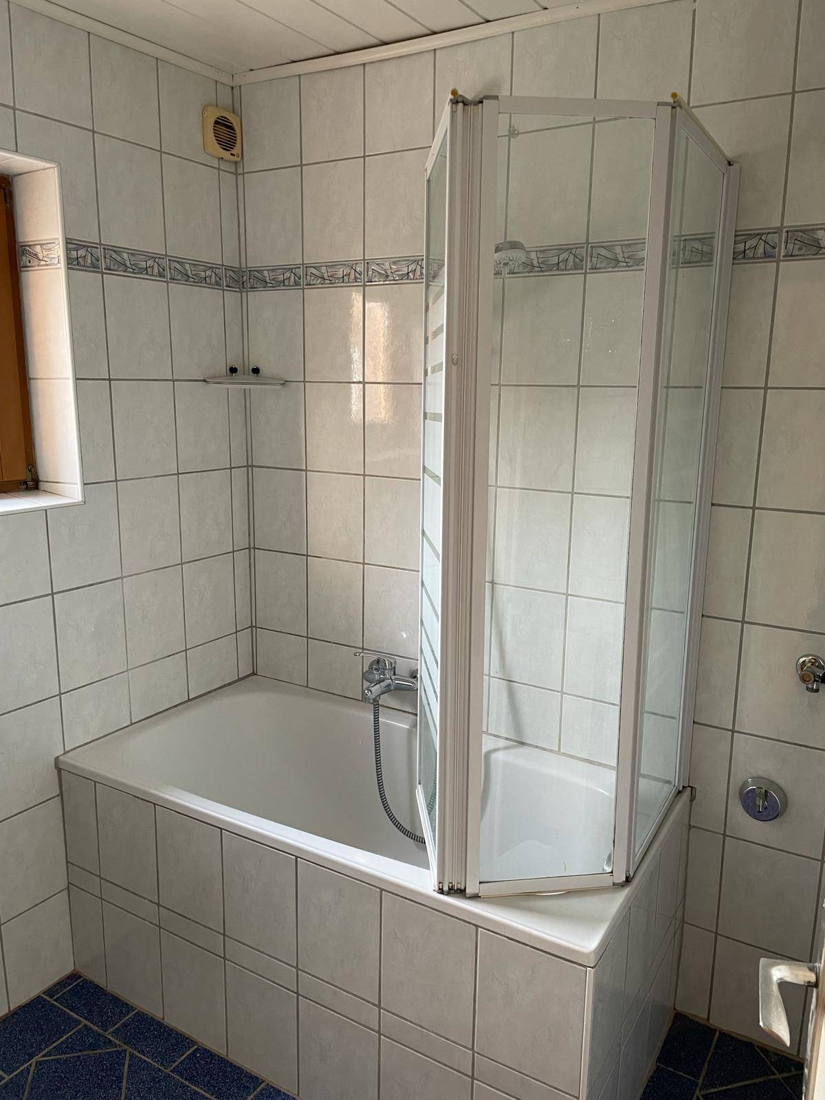 Badezimmer mit Badewanne, Duschabtrennung, Fenster und blau-weißen Fliesen.