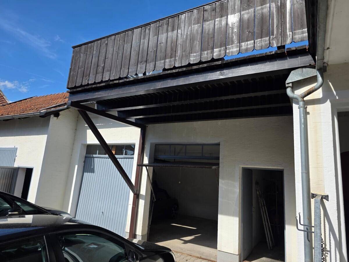 Außenansicht einer Garage mit offenem Tor und einer darüberliegenden Holzkonstruktion, davor ein geparktes Auto.