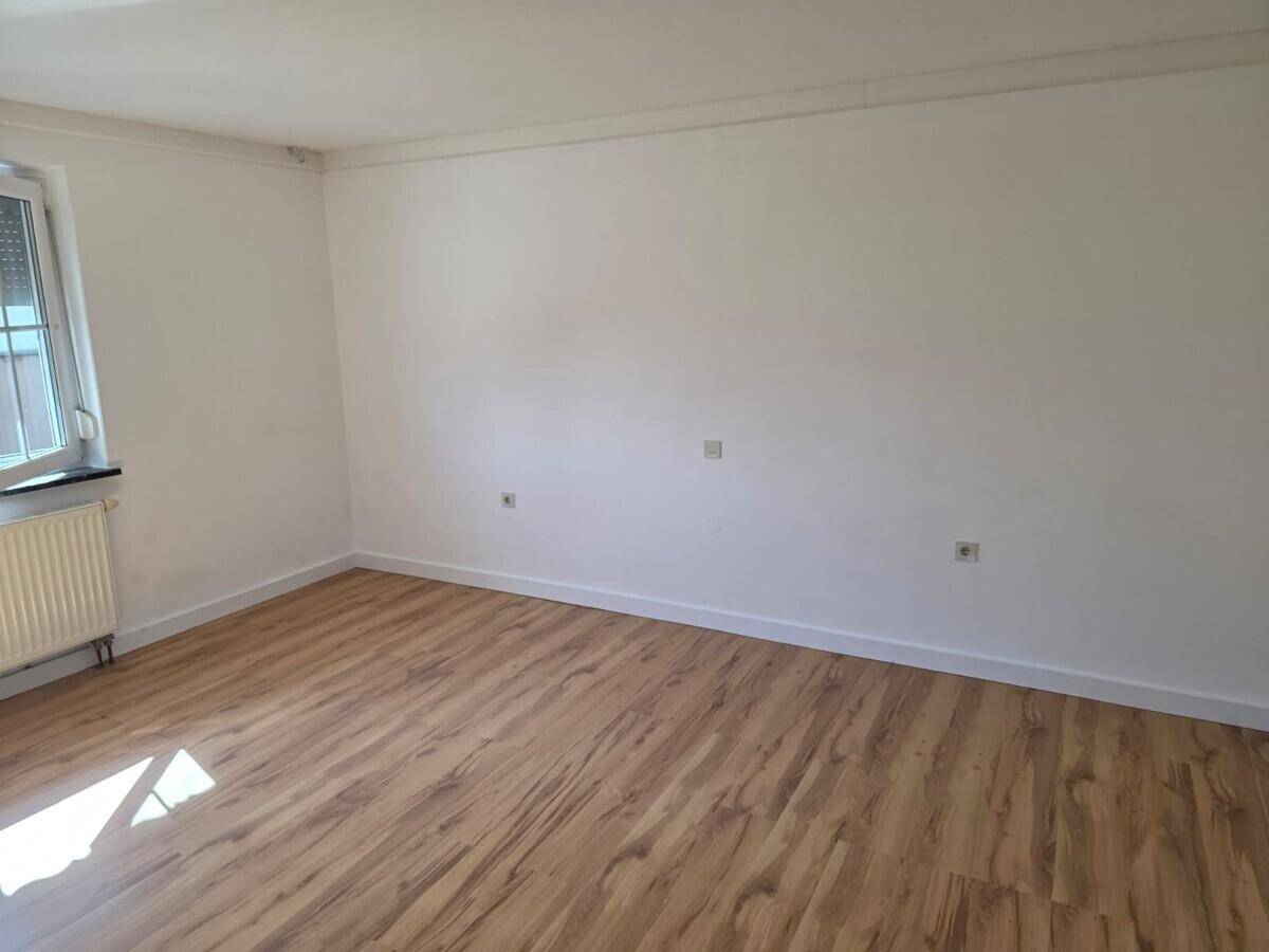 Leerer Raum mit Laminatboden, weißen Wänden und einem Fenster mit Rollladen.