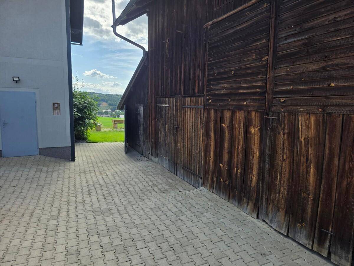 Außenansicht einer Holzscheune neben einem gepflasterten Bereich und einem modernen Gebäude.