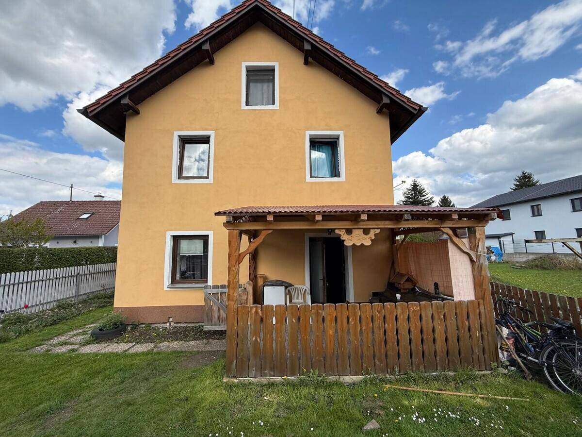 Rückseite des Hauses mit überdachtem Sitzbereich und grünem Garten unter blauem Himmel.